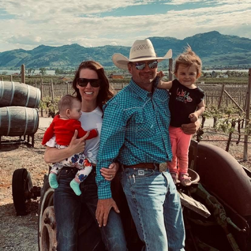 Une famille souriante de quatre personnes, dont deux jeunes enfants, pose en plein air sur un tracteur d'époque, avec des montagnes, des vignobles et de grands tonneaux en bois en arrière-plan.