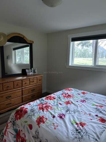 La chambre à coucher confortable est ornée d'un couvre-lit à fleurs, d'une commode en bois avec un miroir, de photos encadrées, d'un chapeau et d'une grande fenêtre offrant une vue pittoresque sur les arbres à l'extérieur.