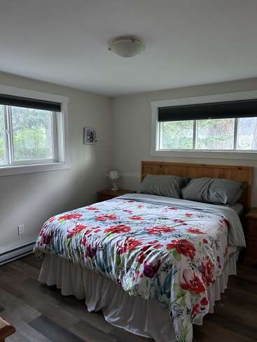 Cette chambre à coucher confortable comprend un lit en bois avec des oreillers gris, une couette à fleurs colorée, deux fenêtres avec des stores, des œuvres d'art et une table de nuit en bois. La lumière naturelle éclaire la pièce.