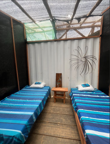 Das Bild zeigt ein schlichtes Zimmer mit zwei Einzelbetten, blau gestreiften Bettüberwürfen, Holzboden und -decke, einem kleinen Tisch und dekorativen Wandbildern.