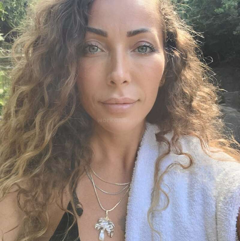 Eine Frau mit langen Locken und klaren Augen posiert im Freien, mit mehreren Halsketten und einem weißen Handtuch über der Schulter, umgeben von viel Grün.