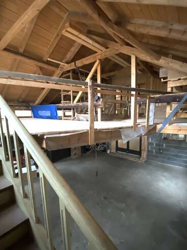 Esta imagen muestra el interior de un loft de madera sin terminar, con vigas a la vista, una escalera, barandillas a medio construir, suelo de hormigón y materiales de construcción esparcidos por todo el espacio.