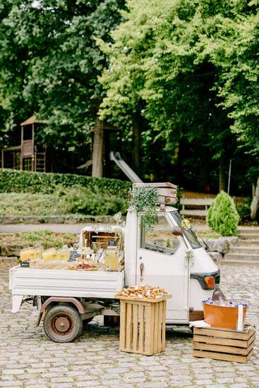 Hay un pequeño food truck blanco instalado al aire libre, que ofrece bebidas y aperitivos sobre cajas de madera, rodeado de vegetación y árboles en un encantador entorno de parque.