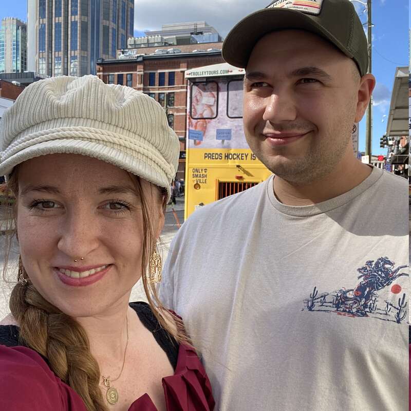 A smiling woman and man pose together outdoors in a city. Tall buildings, a yellow tour bus, and blue sky with clouds are visible in the background.