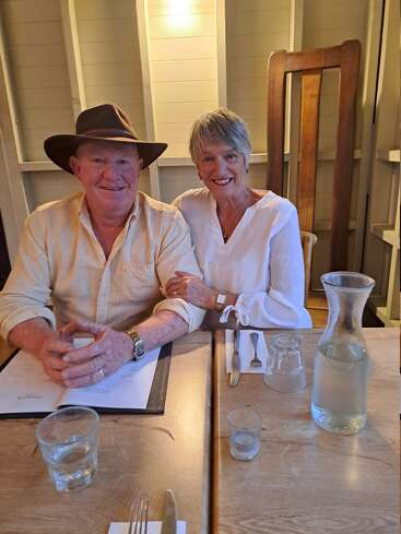 Una pareja de ancianos se sientan juntos en la mesa de un restaurante, sonriendo afectuosamente. El hombre lleva sombrero y camisa de rayas; la mujer viste de blanco. Hay agua y menús.