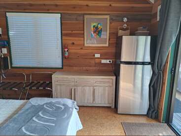 Esta acogedora habitación de madera cuenta con cama, cómoda, portaequipajes, frigorífico, ventana con persianas, cortinas, pintura de colores, extintor e iluminación natural.