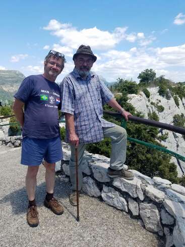 Dois homens posam juntos ao ar livre num mirante panorâmico de pedra. Um deles está vestindo camiseta e bermuda, o outro, um chapéu e camisa xadrez, segurando uma bengala.
