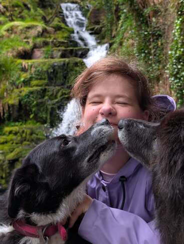 Dos perros lamen cariñosamente a una alegre persona con chaqueta morada, enmarcada por una exuberante vegetación y una cascada al fondo.