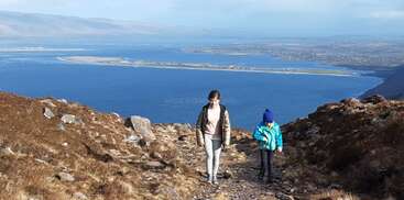 Dos personas suben por un sendero rocoso en una montaña con un impresionante lago azul y tierras lejanas visibles bajo un cielo claro y brillante.