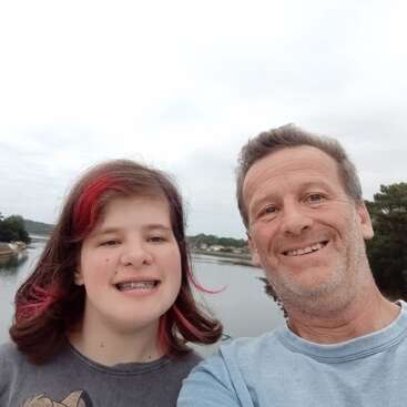 Un homme souriant et une fille avec des mèches roses et un appareil dentaire prennent un selfie joyeux en plein air, près d'une rivière calme, d'arbres et d'un ciel nuageux.