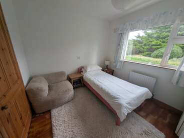 Se trata de un dormitorio pequeño y acogedor con una cama individual, sillón, mesilla de noche, lámpara, alfombra mullida, suelo de madera, ventana grande y vistas verdes al exterior.