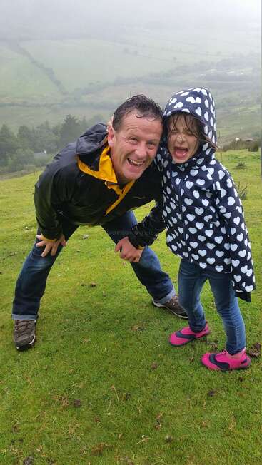 Ein Mann und ein junges Mädchen, beide fröhlich lächelnd in Regenjacken, posieren zusammen auf einem üppigen, grünen, nebligen Hügel mit malerischer Landschaft im Hintergrund.