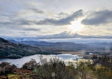 Eine ruhige Landschaft mit einem spiegelnden See, kahlen Bäumen, sanften Hügeln und schneebedeckten Bergen unter einem wolkenverhangenen Himmel, der von der durchscheinenden Sonne sanft beleuchtet wird.