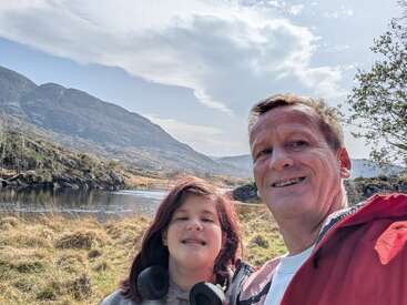Ein Mann und ein junges Mädchen lächeln für ein Selfie im Freien an einem Fluss, mit malerischen Bergen, grasbewachsenen Feldern und einem teilweise bewölkten Himmel im Hintergrund.