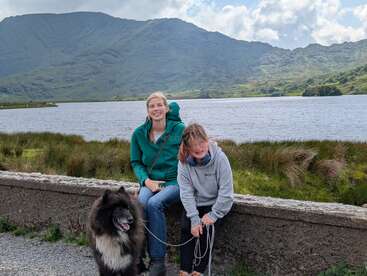 Zwei Menschen und ein flauschiger schwarzer Hund sitzen lächelnd auf einer Steinmauer neben einem malerischen See mit grünen Bergen und einem bewölkten Himmel im Hintergrund.