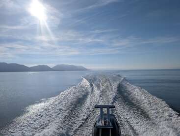 Ein Boot rast bei strahlendem Sonnenschein über das ruhige, blaue Wasser und hinterlässt ein schäumendes Kielwasser. In der Ferne erheben sich die Berge am Horizont unter einem klaren Himmel.