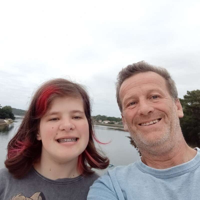 Un hombre sonriente y una niña con mechas rosas y aparatos dentales se toman un alegre selfie al aire libre, cerca de un río tranquilo, árboles y cielo nublado de fondo.
