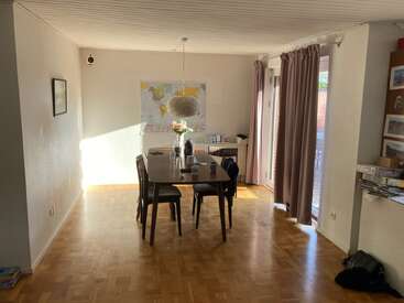 Un luminoso comedor con una mesa de madera, seis sillas, un centro de mesa con un jarrón de flores, un mapamundi en la pared y la luz del sol que entra por grandes ventanales con cortinas.
