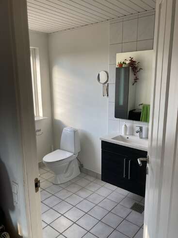 Un cuarto de baño limpio y moderno con suelo de baldosas blancas, paredes blancas, inodoro, tocador negro, lavabo, espejo, pequeñas plantas y una toalla colgada junto al lavabo.