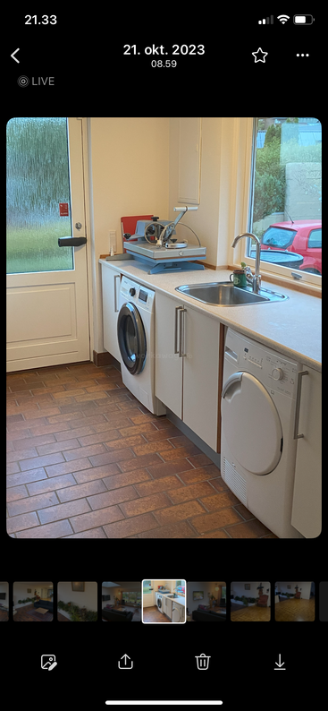 Esta imagen muestra un lavadero ordenado con lavadora, secadora, encimera, fregadero, cortadora grande, ventana, puerta y suelo de baldosas marrones. Iluminación brillante.