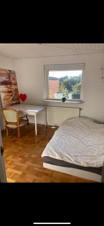 Un dormitorio luminoso y sencillo con suelo de madera, paredes blancas, cama, escritorio, silla, cuadro de colores, decoración de corazones rojos y una ventana con vistas.