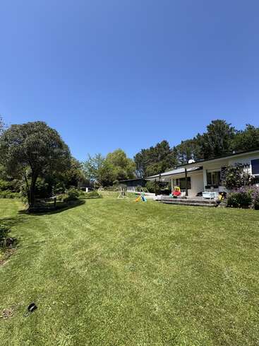 Um espaçoso quintal verde com grama recém-cortada, cercado por árvores, uma casa branca, equipamentos de recreação infantil, uma varanda e um céu azul claro e brilhante.