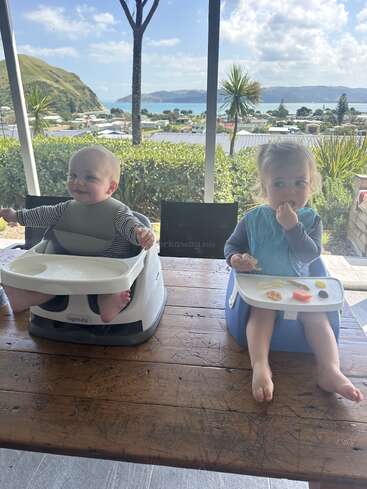 Duas crianças pequenas sentam-se em assentos elevatórios em uma mesa de madeira, comendo lanches. Atrás delas, há uma bela vista panorâmica de montanhas, árvores e do oceano.