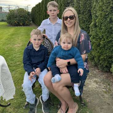 The image depicts a woman sitting on a chair with two young boys, one on her lap and the other beside her, in a grassy area with a hedge wall and cloudy sky in the background.