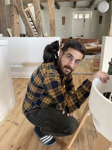 A man in glasses and a plaid shirt crouches indoors. A small black kitten sits on his shoulder. The room has rustic wooden beams and cozy décor.