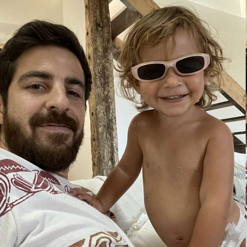 A bearded man and a smiling child with messy hair and sunglasses take a cute, playful selfie indoors, capturing a happy, relaxed family moment together.