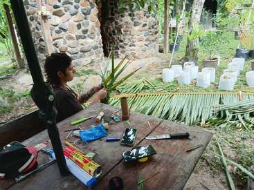 Une personne est assise à l'extérieur, près d'un bâtiment rustique en pierre, et tisse des feuilles de palmier. La table est encombrée d'outils, d'un téléphone, de matériaux d'artisanat et de divers objets.