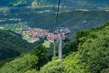 A picturesque cable car ascends over lush green forested mountains, offering a breathtaking view of a quaint village nestled in a scenic, fertile valley below.