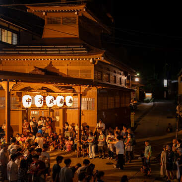 A lively crowd gathers at night outside a traditional Japanese wooden building, warmly lit by lanterns, enjoying a festival or community event, with excitement and anticipation.