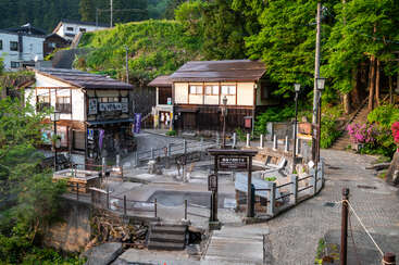 This image shows a quaint Japanese village scene with traditional wooden buildings, a steaming public hot spring, lush greenery, stone paths, flowers, and peaceful morning sunlight.