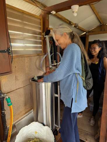 Una mujer mayor con un jersey azul maneja una máquina metálica en una habitación de estilo rústico. Detrás de ella hay otra mujer con gafas, que observa el proceso con una sonrisa.