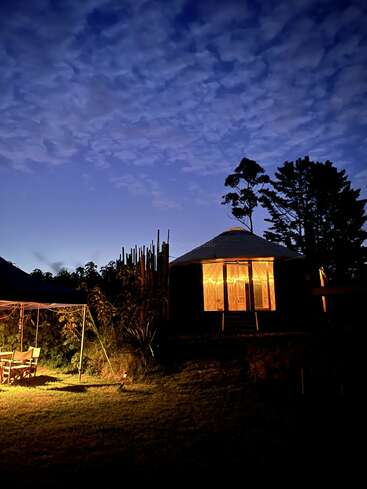Une yourte chaleureuse, baignée d'une douce lumière la nuit, se dresse au milieu des arbres sous un ciel d'un bleu profond. Des lumières tamisées éclairent un coin repas en plein air tout proche, créant une atmosphère paisible.