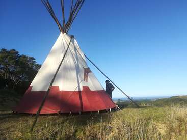 Un grand tipi se dresse sur une colline verdoyante, sous un ciel bleu limpide. Une personne coiffée d'un chapeau se tient à côté, le regard tourné vers les arbres et l'océan au loin.