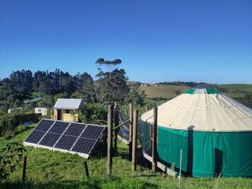 Une yourte à la base verte se dresse à côté de panneaux solaires dans un cadre rural verdoyant, entourée d'arbres, de collines, de constructions en bois et d'un ciel bleu limpide.