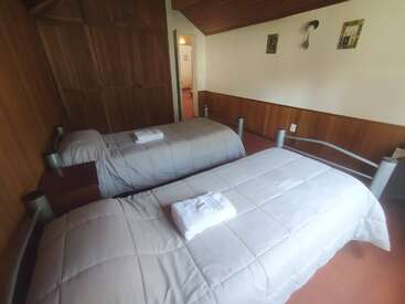 This image shows a cozy bedroom with two single beds, each made neatly with gray comforters and towels on top. Wooden walls and simple decor.