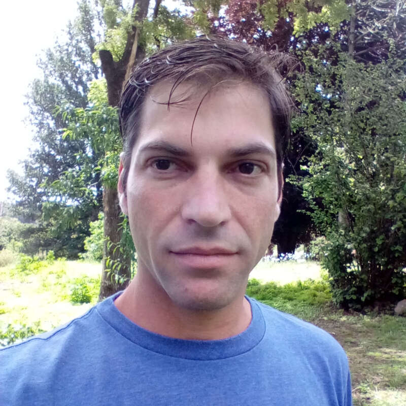 The image depicts a man standing outdoors, wearing a blue shirt, with trees and grass visible in the background on a sunny day.