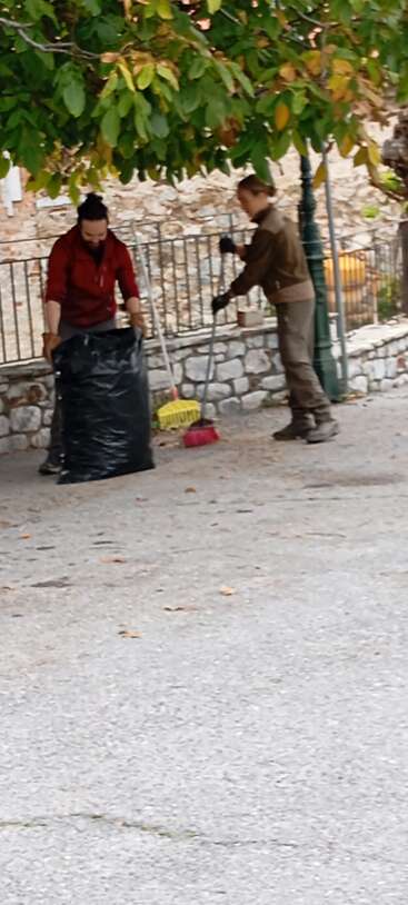Two people clean outdoors under a tree. One sweeps leaves into a dustpan, the other holds a large black trash bag. Stone wall and fence behind.