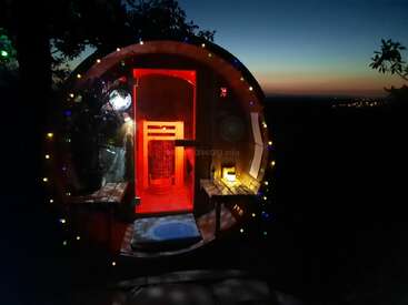 Uma cabana aconchegante em forma de barril brilha com luzes vermelhas e amarelas ao anoitecer, decorada com luzes coloridas de fadas, aninhada ao ar livre sob um céu escuro, convidando ao relaxamento.