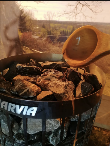 A wooden ladle pours water onto hot sauna stones inside a metal heater, creating steam. The background shows a scenic outdoor landscape through a window.