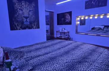 A bedroom illuminated by blue lights features leopard-print bedding, large feline-themed artwork, a wall mirror with bulbs, a drink cart, and champagne with glasses.