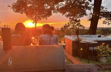 Two people sit together at an outdoor table during sunset, surrounded by trees, string lights, a hot tub, and a cozy, relaxing atmosphere.