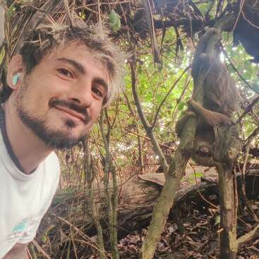 A man smiling takes a selfie in a lush forest, while a sloth hangs upside down from a tree branch behind him among dense greenery.