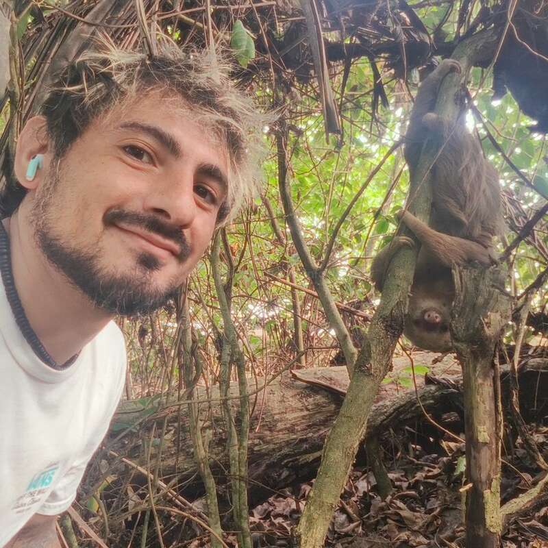 A man smiling takes a selfie in a lush forest, while a sloth hangs upside down from a tree branch behind him among dense greenery.