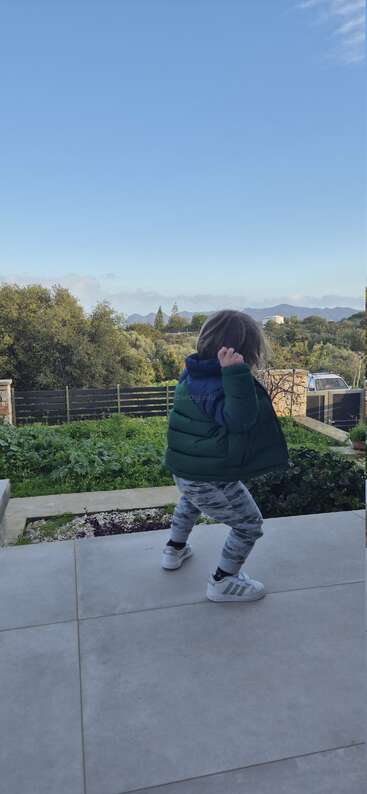 A young child in a green jacket and patterned pants stands on a patio, backed by lush greenery, trees, mountains, and a clear blue sky.
