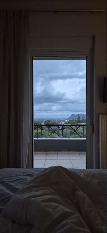 A cozy bed overlooks an open balcony door, with cloudy skies and a scenic mountain view framed by curtains, evoking calm, relaxation, and peaceful morning vibes.