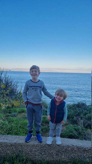 Two smiling young boys stand on a low ledge, with lush greenery and the expansive sea behind them. The sky is clear and blue, serene.